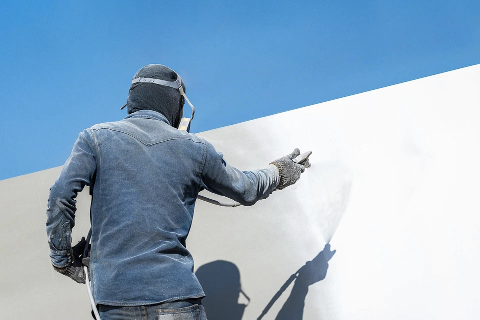 Gros plan professionnel montrant un artisan en vêtements de travail appliquant un isolant thermique (laine minérale, polystyrène ou ouate) sur une paroi intérieure ou un mur, avec équipements et matériaux d'isolation visibles. L'image doit montrer clairement un travail d'isolation (non de peinture) dans un contexte de rénovation résidentielle, reflétant l'expertise technique et professionnelle requise pour les travaux d'isolation RGE.